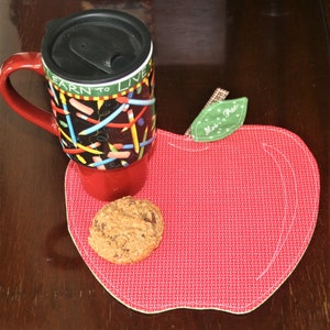 May include: Red apple-shaped coaster with a green leaf that says "Miss Pat" on it. A red travel mug with a black lid and colorful pencils printed on it is sitting on the coaster. A chocolate chip cookie is also on the coaster.