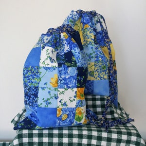 May include: Two blue and yellow drawstring bags made of patchwork fabric with floral patterns. The bags are sitting on a green and white checkered tablecloth.