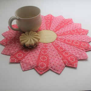May include: A pink and white fabric coaster shaped like a flower with a scalloped edge. The coaster has a yellow center and is perfect for protecting surfaces from hot drinks.