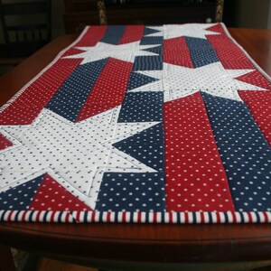 May include: A red, white, and blue table runner with white stars on a blue and red background. The runner has a white and red striped border.