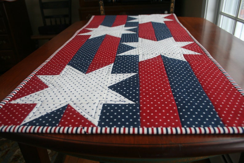 May include: A red, white, and blue table runner with white stars on a blue and red background. The runner has a white and red striped border.