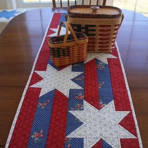 May include: A red, white, and blue quilted table runner with a star pattern. Two wicker baskets with red, white, and blue woven designs are on top of the runner.