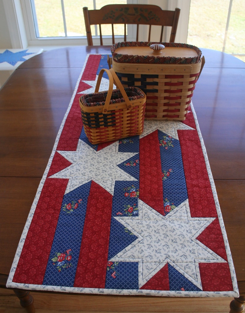 May include: A red, white, and blue quilted table runner with a star pattern. Two wicker baskets with red, white, and blue woven designs are on top of the runner.