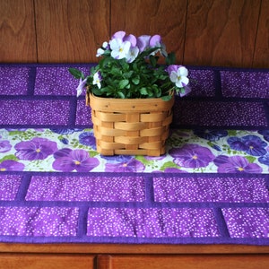 Puede incluir: Un camino de mesa decorativo morado con un diseño de ladrillos y un diseño floral. Una cesta tejida contiene una planta en maceta con pensamientos morados y blancos. El camino de mesa está sobre una superficie de madera.