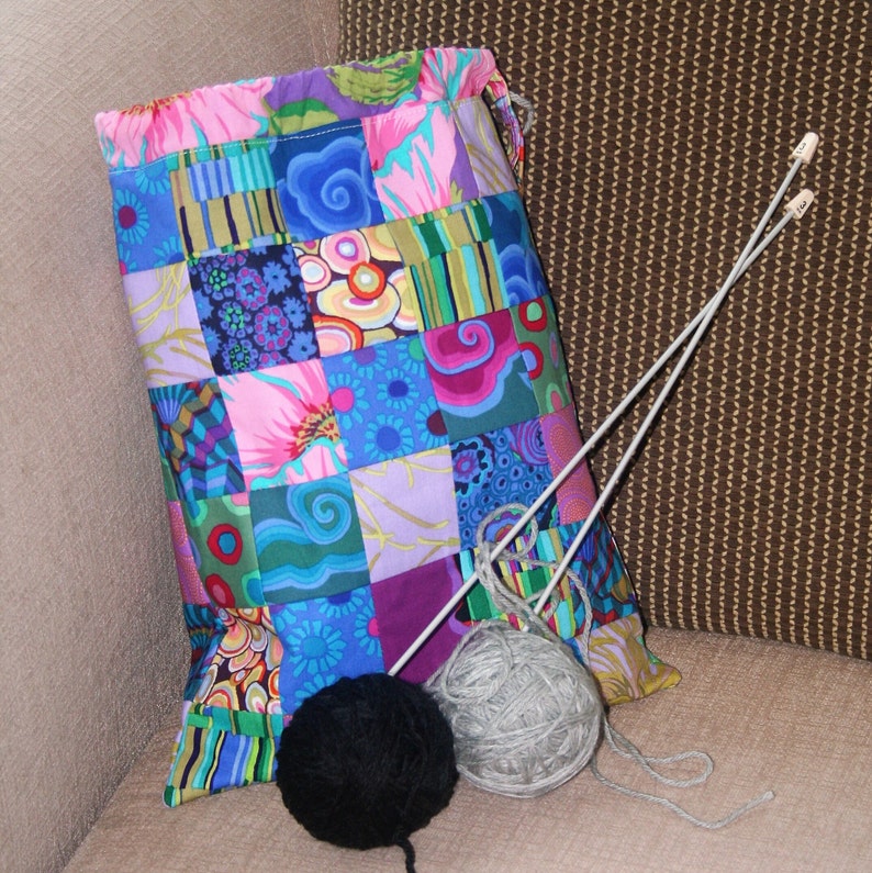 May include: A patchwork fabric drawstring bag with a colorful design. The bag is filled with yarn and knitting needles.