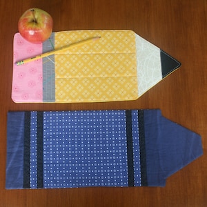 May include: A yellow pencil-shaped fabric book cover with a pink, gray, and white patterned design. The cover is lying on a wooden surface. A blue fabric book cover with white polka dots and black stripes is also shown.