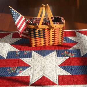 May include: A red, white, and blue quilted table runner with a woven basket and an American flag. The runner features a star design and is perfect for a patriotic themed table setting.