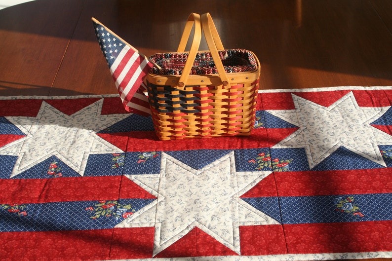 May include: A red, white, and blue quilted table runner with a woven basket and an American flag. The runner features a star design and is perfect for a patriotic themed table setting.