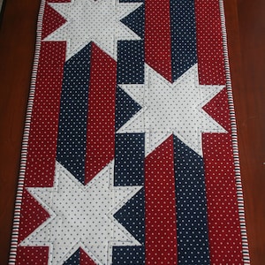 May include: A red, white, and blue table runner with a patriotic star design. The runner is made of fabric with white polka dots on a red and blue background.