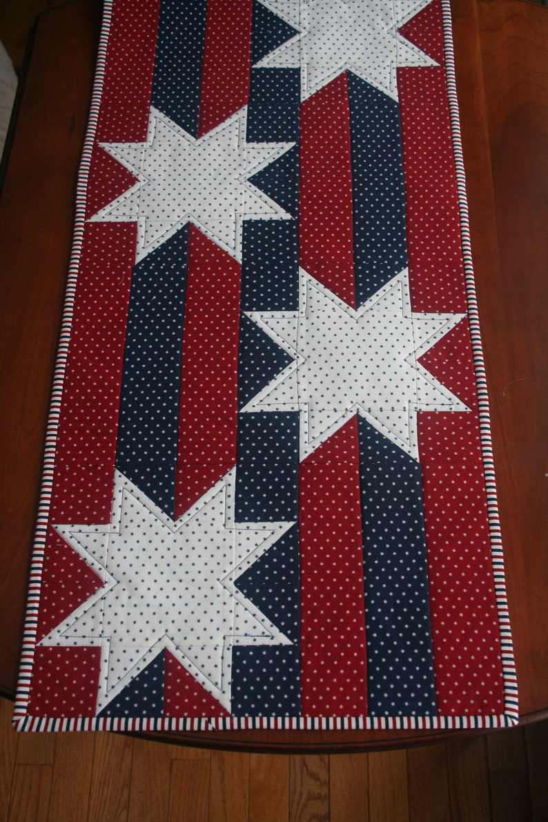 May include: A red, white, and blue table runner with a patriotic star design. The runner is made of fabric with white polka dots on a red and blue background.