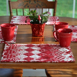 May include: Four red mugs sit on a wooden table with red and white quilted placemats. A red pot with a green plant sits in the center of the table.
