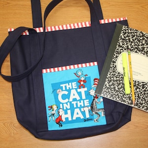 Pode incluir: Uma bolsa tote de lona azul com listras vermelhas e brancas. A bolsa tem um bolso na frente com um gráfico dos personagens do livro "O Gato no Chapéu" de Dr. Seuss.