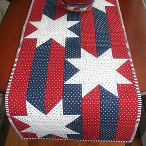 May include: Red, white, and blue table runner with white stars on a red and blue striped background. The runner has a white and red trim.