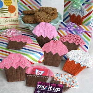 Puede incluir: Una colección de tarjeteros y artículos decorativos hechos a mano con forma de cupcake. Los cupcakes presentan diseños de glaseado rosa, naranja y blanco con envoltorios marrones. También hay un plato de galletas y regalos envueltos con lazos.