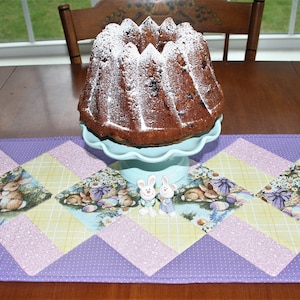 Può includere: Un runner da tavolo trapuntato viola, rosa e giallo con un motivo a quadri e disegni di coniglietti. Il runner è su un tavolo di legno con una torta su un piedistallo blu al centro.