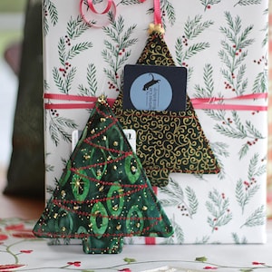 Peut inclure: Deux décorations de Noël en forme de sapin en tissu vert avec des accents dorés et rouges. Les décorations sont suspendues sur un fond blanc avec un motif vert et rouge.