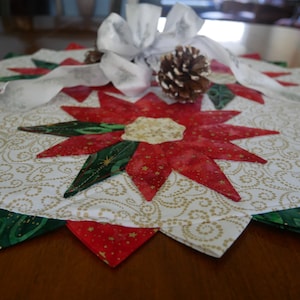 Peut inclure: Un chemin de table blanc avec un motif de spirales dorées et un design de poinsettia rouge et vert. Le chemin de table est décoré d'un nœud blanc et d'une pomme de pin.