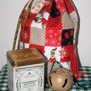 May include: A red and green patchwork fabric gift bag with a drawstring closure. The bag is filled with a tin of Harney & Sons Gingerbread Festival black tea with ginger, cinnamon, and molasses. A brown jingle bell with a green ribbon is also included.