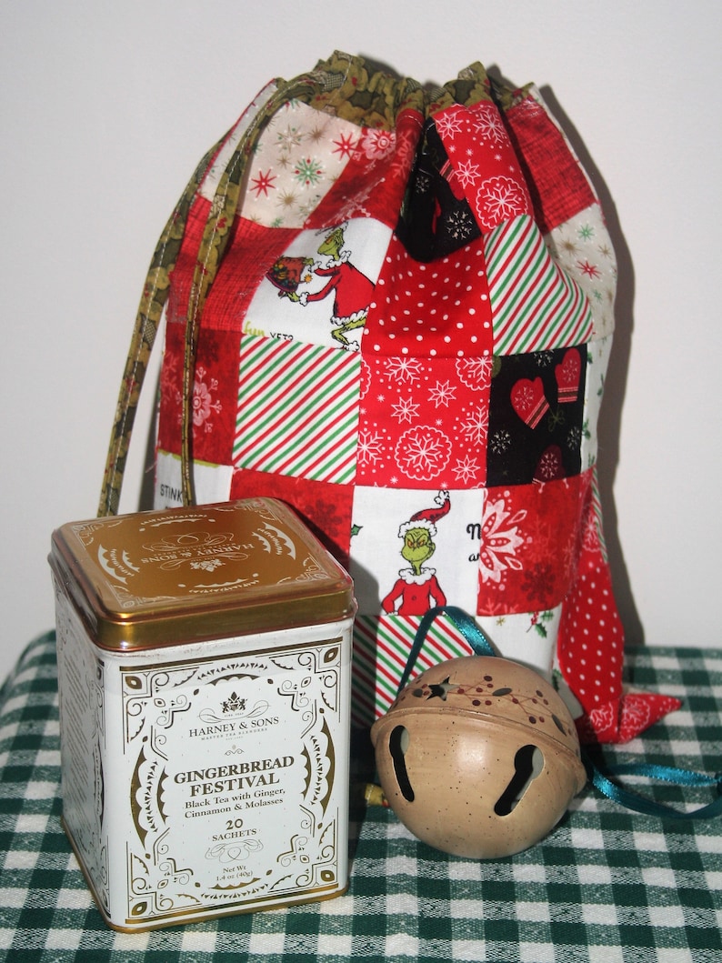 May include: A red and green patchwork fabric gift bag with a drawstring closure. The bag is filled with a tin of Harney & Sons Gingerbread Festival black tea with ginger, cinnamon, and molasses. A brown jingle bell with a green ribbon is also included.