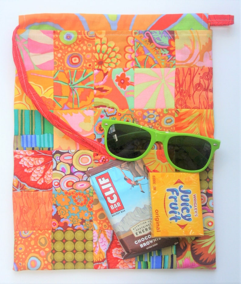 May include: A patchwork fabric drawstring bag with a red strap, green sunglasses, a Clif Bar energy bar, and a Juicy Fruit gum pack. The bag is made of colorful fabric with a variety of patterns.