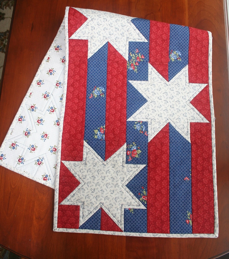May include: A red, white, and blue table runner with a star pattern. The runner is made of fabric with a floral print and is perfect for a patriotic themed party.