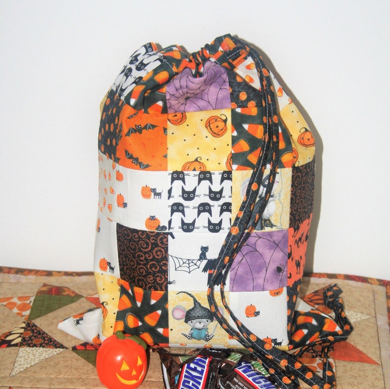 May include: A Halloween-themed drawstring bag made of patchwork fabric with various designs, including pumpkins, bats, cats, candy corn, and spiders. The bag is purple, orange, black, and white.