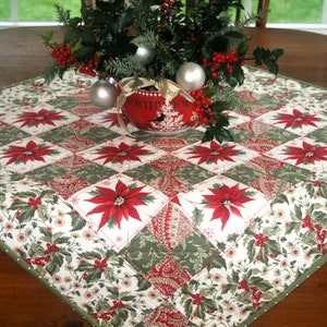 Könnte beinhalten: Ein weißer und grüner gesteppter Tischläufer mit einem roten Weihnachtsstern-Design. Der Läufer ist mit einem Mittelstück aus Stechpalme und roten Beeren in einem roten und weißen Keramiktopf mit der Aufschrift "Nordpol" verziert.