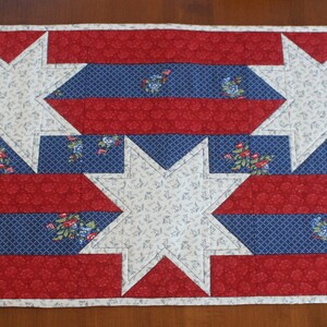 May include: A red, white, and blue quilted table runner with a star pattern. The runner is made of fabric with a floral print.