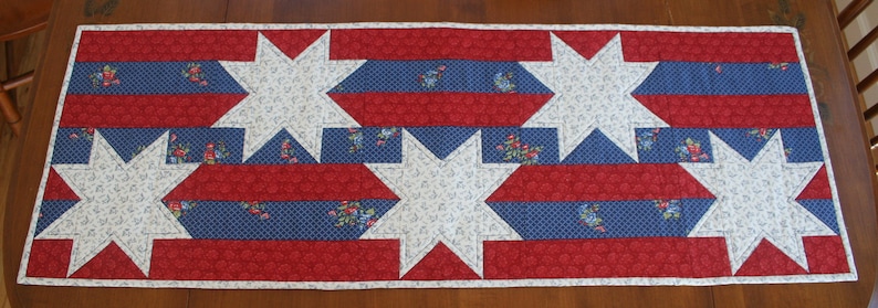 May include: A red, white, and blue quilted table runner with a star pattern. The runner is made of fabric with a floral print.