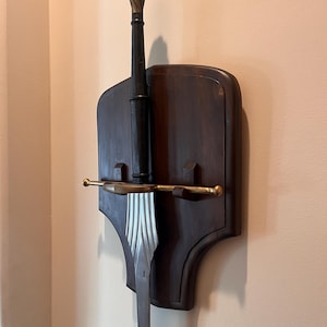 Könnte beinhalten: Ein dekoratives Schwert und ein Ausstellungsständer. Das Schwert hat eine silberne Klinge und einen dunklen Griff mit einem goldfarbenen, spiralförmigen Knauf. Das Schwert ist auf einer dunkelbraunen Holztafel mit Messingakzenten montiert.