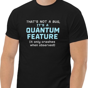 May include: Black t-shirt featuring the text "THAT'S NOT A BUG, IT'S A QUANTUM FEATURE (it only crashes when observed)" in white and light blue. The shirt has a crew neck and short sleeves.