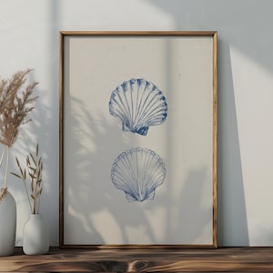 Op de afbeelding: Een ingelijste print met twee blauwe schelpenillustraties op een crèmekleurige achtergrond. Het kunstwerk wordt gepresenteerd op een houten oppervlak, samen met witte vazen met gedroogde planten en een decoratieve vaas. Thema: kustdecoratie.