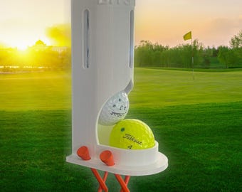 May include: A white golf ball dispenser with the word "BITS" on top. The dispenser holds two golf balls, one white and one yellow. Two orange golf tees are attached to the bottom. The background shows a golf course with a flag.