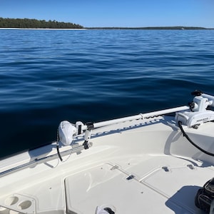 Puede incluir: Un barco blanco con un motor de pesca el&eacute;ctrico blanco montado en la proa. El barco est&aacute; en una masa de agua con una l&iacute;nea de costa en la distancia.