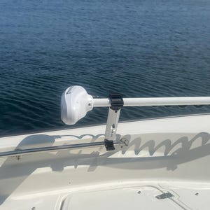Puede incluir: Un motor de pesca el&eacute;ctrico blanco montado en un barco blanco con una varilla de acero inoxidable y abrazaderas negras. El motor est&aacute; montado en el lateral del barco y est&aacute; orientado hacia adelante. El barco est&aacute; en el agua y hay una costa verde en el fondo.