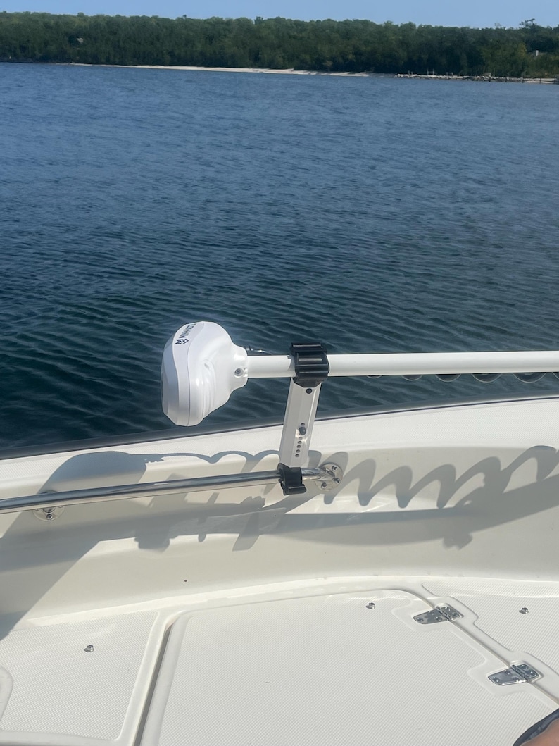 Puede incluir: Un motor de pesca el&eacute;ctrico blanco montado en un barco blanco con una varilla de acero inoxidable y abrazaderas negras. El motor est&aacute; montado en el lateral del barco y est&aacute; orientado hacia adelante. El barco est&aacute; en el agua y hay una costa verde en el fondo.