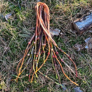 Op de afbeelding: Een set kleurrijke dreadlock-extensies met kralen. De extensies zijn gemaakt van wol en hebben een verscheidenheid aan kleuren, waaronder oranje, geel, rood en bruin. De kralen zijn klein en rond en zijn gelijkmatig verdeeld over de extensies.