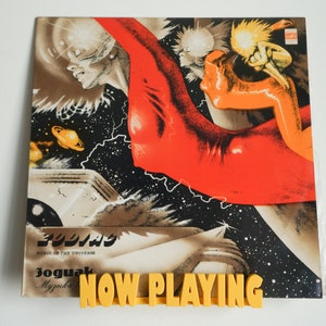 May include: A vintage vinyl record cover with a colourful abstract design featuring a red, orange and yellow figure against a black background with white stars. The text "Joguak Muzyka" and "Music in the Universe" are printed on the cover. The words "NOW PLAYING" are printed in yellow on a black background.