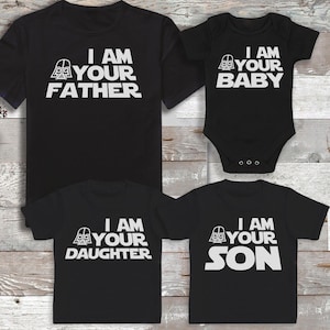 Peut inclure: Des t-shirts noirs et un body de b&eacute;b&eacute; avec du texte blanc qui dit "I AM YOUR FATHER", "I AM YOUR BABY", "I AM YOUR DAUGHTER" et "I AM YOUR SON". Les chemises pr&eacute;sentent un graphisme de casque de Dark Vador.
