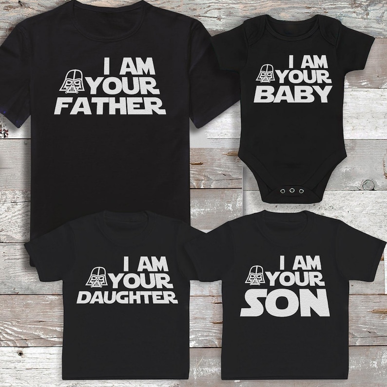 Peut inclure: Des t-shirts noirs et un body de b&eacute;b&eacute; avec du texte blanc qui dit "I AM YOUR FATHER", "I AM YOUR BABY", "I AM YOUR DAUGHTER" et "I AM YOUR SON". Les chemises pr&eacute;sentent un graphisme de casque de Dark Vador.