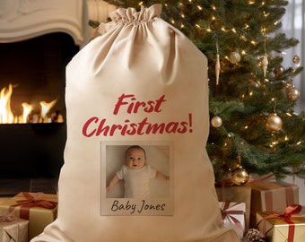 May include: A beige canvas Santa sack with the text "First Christmas!" in red. A photo of a baby is in a white frame with the text "Baby Jones" below the photo.