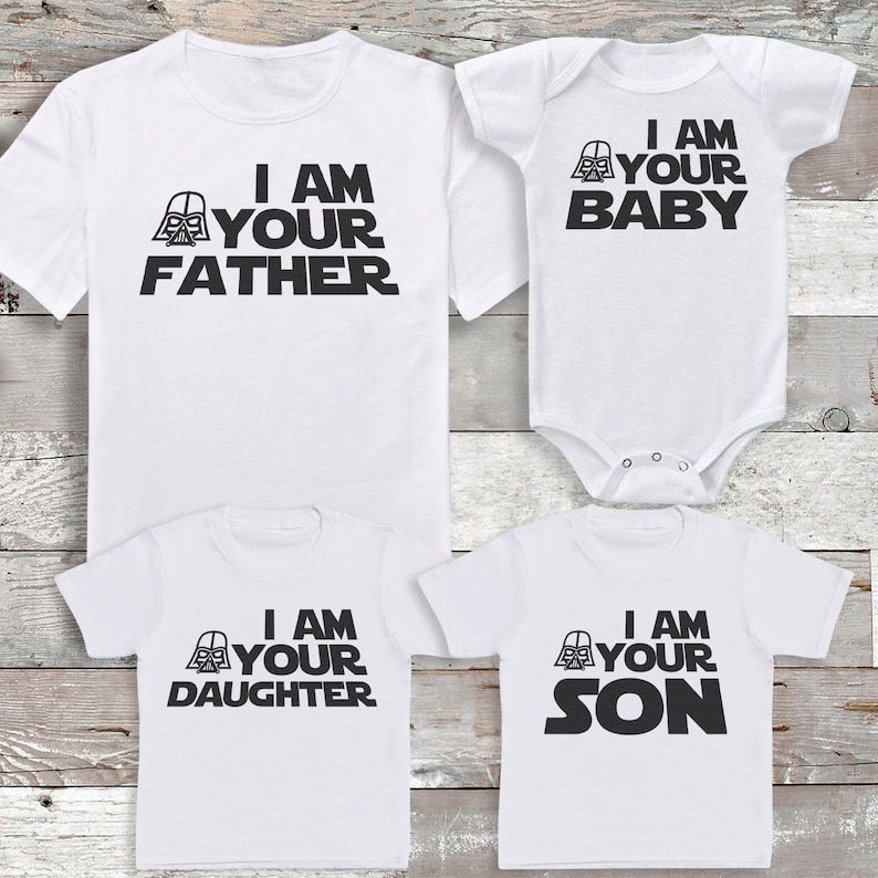 Peut inclure: Quatre t-shirts blancs et un body pour b&eacute;b&eacute; avec du texte noir. Les chemises indiquent "I AM YOUR FATHER", "I AM YOUR DAUGHTER" et "I AM YOUR SON". Le body dit "I AM YOUR BABY". Chaque article pr&eacute;sente un graphisme de casque de Dark Vador.