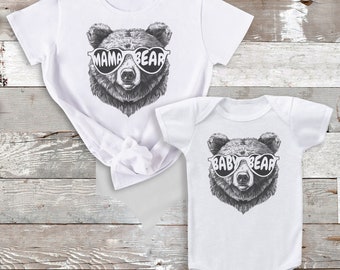 May include: White t-shirt with a black and white illustration of a bear wearing heart-shaped sunglasses and the text "Mama Bear". A white bodysuit with a black and white illustration of a bear wearing heart-shaped sunglasses and the text "Baby Bear".