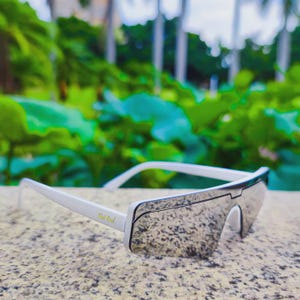 May include: White-framed sunglasses with mirrored lenses. The sunglasses have a modern, angular design with a sleek, minimalist aesthetic. The text "Real Kool" is printed on the left arm of the sunglasses. The background is blurred green foliage.