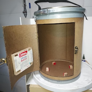 Puede incluir: Un contenedor cilíndrico con una puerta con bisagras, construido con cartón y metal. El contenedor tiene una parte superior e inferior de metal plateado, con un asa negra. Una caja de Coleman Silk-lite Mantles está adherida a la puerta. En el interior hay varios bloques de madera pequeños.