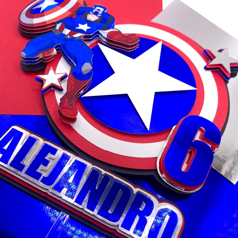 Puede incluir: Adorno para tarta en capas con tem&aacute;tica de Capit&aacute;n Am&eacute;rica, en colores azul, rojo y blanco. El dise&ntilde;o incluye una figura del Capit&aacute;n Am&eacute;rica, un escudo, estrellas, el n&uacute;mero 6 y el nombre "Alejandro".