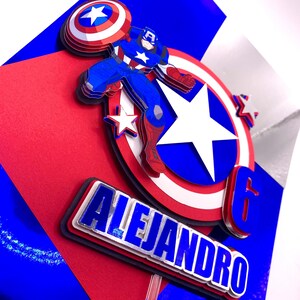 Puede incluir: Un adorno para tarta de Capit&aacute;n Am&eacute;rica en capas con el nombre "Alejandro" y el n&uacute;mero "6". El dise&ntilde;o presenta al Capit&aacute;n Am&eacute;rica, un escudo y estrellas en rojo, blanco y azul. El fondo es rojo y azul.