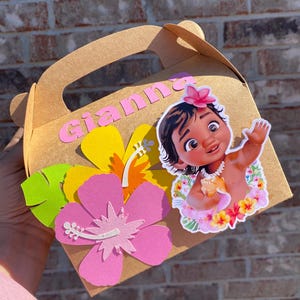 May include: A brown cardboard gift box with a handle, decorated with a pink flower, a yellow flower, and a cartoon image of a baby girl. The name "Gianna" is written in pink letters on the box.