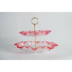 May include: Two-tiered glass serving stand with a gold metal handle. The stand has a scalloped edge and a pink and white ombre design.