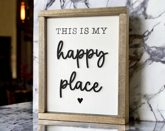 May include: A framed sign with the words "THIS IS MY happy place" in black lettering on a white background. The frame is a light brown color, and a small black heart is below the word "place".